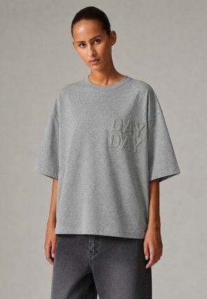 Kvinde iført en løs grå T-shirt med hævede "DAY DAY" bogstaver og mørkegrå bukser mod en ensfarvet baggrund.