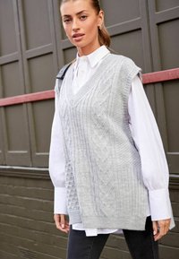 Gilet tricoté gris clair avec un col en V, présentant des motifs en torsades et un ourlet en côtes. Porté par-dessus une chemise blanche à col, associé à un jean foncé.