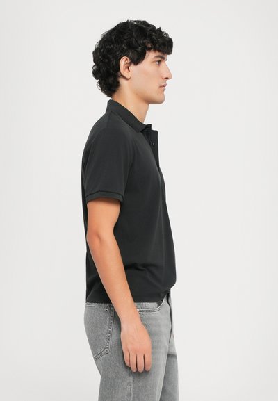 Polo noir à manches courtes, avec col et patte de boutonnage. Associé à un jean gris clair. Le modèle se tient de profil sur un fond blanc.