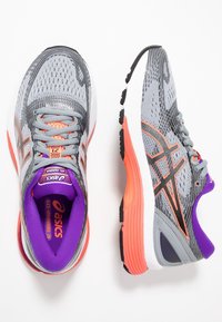 Zapatillas de correr grises con acentos naranjas, parte superior de malla transpirable, suela de goma negra resistente y un interior púrpura. Presenta un logo distintivo y cordones.