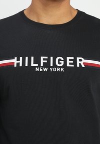 Czarny t-shirt z okrągłym dekoltem z napisem "HILFIGER NEW YORK" na klatce piersiowej, podkreślony białymi i czerwonymi poziomymi paskami.