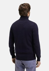 Homme aux cheveux bruns portant un pull à col roulé bleu marine et un pantalon bleu clair, vu de dos sur un fond uni.