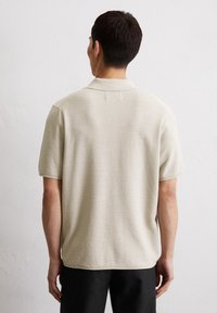 Beige short-sleeve knit polo shirt with a textured weave and a classic collar, featuring subtle seam detailing at the back.