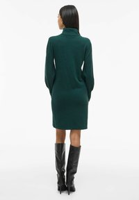 Vestido verde de manga larga con cuello alto, que presenta una silueta ajustada y una textura suave, combinado con botas de cuero negras hasta la rodilla.