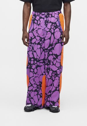 Pantalon ample avec un motif abstrait violet et noir et des panneaux latéraux en mesh orange, porté avec des chaussures noires et une chemise noire.
