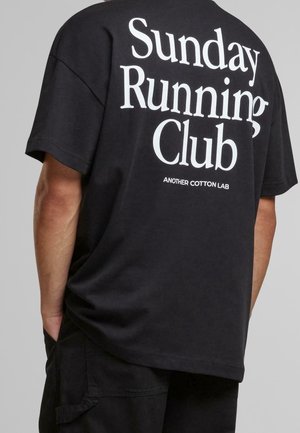 T-shirt noir avec texte blanc "Sunday Running Club" et texte plus petit "Another Cotton Lab" dans le dos, porté par une personne les mains dans les poches.