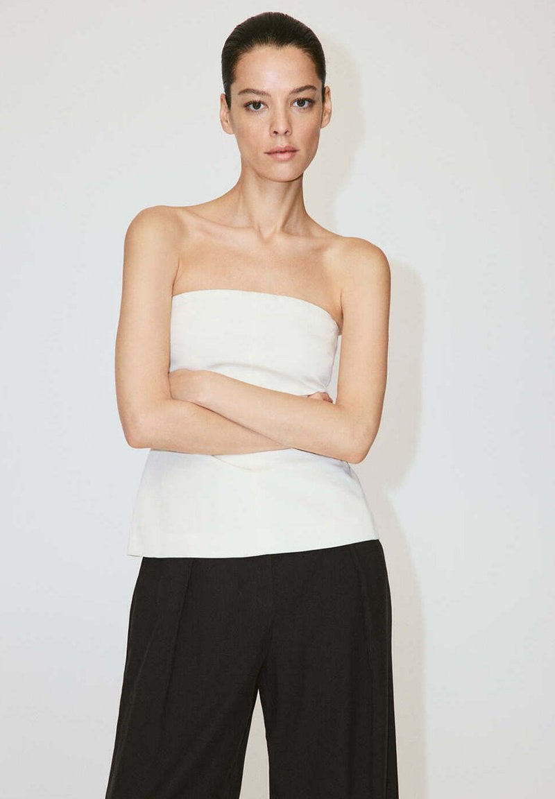 Femme aux cheveux foncés, vêtue d'un haut blanc sans bretelles et d'un pantalon noir, debout les bras croisés devant un fond clair uni.