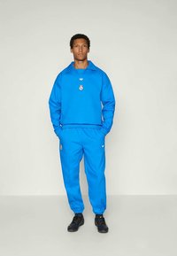 Blå atletisk treningsdress laget av glatt stoff. Har en genser med krage og logo, samt matchende bukser med elastisk midje og sidelommer.