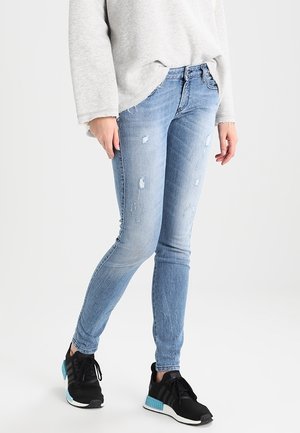 Persoon die lichtblauwe versleten skinny jeans draagt, een grijs shirt met lange mouwen en zwarte sneakers met blauwe accenten, staand tegen een effen achtergrond.