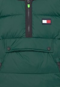 Giacca a vento verde con zip nera, tasca con zip elogo a patch nei colori rosso, bianco e blu. Texture liscia e imbottita.