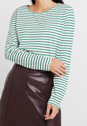 Žena v bílém a zeleném pruhovaném topu s dlouhým rukávem a tmavě hnědé kožené sukni, stojící před jednobarevným pozadím.