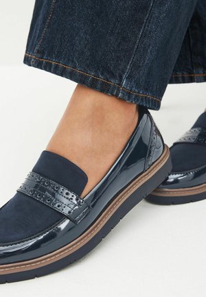 Mocassins bleu marine avec une finition brillante, présentant des accents en daim et une sangle perforée. Semelle épaisse marron offrant confort et soutien.