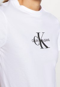 Bílý bavlněný tričko s černě potisknutým logem a textem "CALVIN K JEANS." Kulatý výstřih a krátké rukávy. Hladká textura.