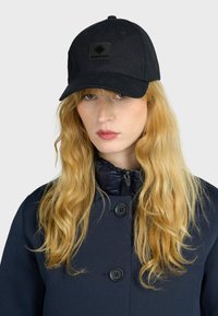 Black cotton baseball cap with a structured crown, flat brim, and a front patch featuring a logo. Worn with a dark coat and a quilted collar.