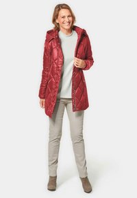 Rode gewatteerde lange jas met capuchon, met een ritssluiting en zijzakken. Het model draagt een lichte top en beige broek, met grijze enkellaarsjes.