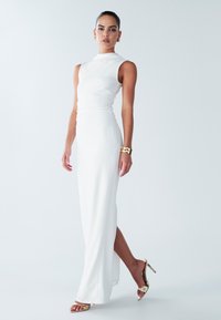 Robe blanche sans manches avec un corsage ajusté et une fente latérale. Tissu lisse avec un léger éclat. Complétée par des sandales à talons dorés.