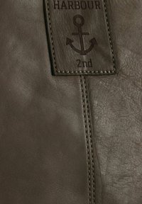 Detalle de bolsa de cuero marrón con una etiqueta cosida que muestra un logo de ancla y las palabras "HARBOUR 2nd." La textura es suave con variaciones sutiles.