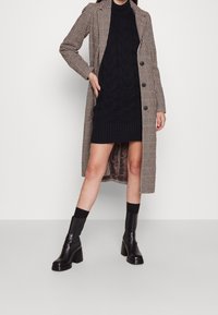 Cappotto a scacchi marrone e nero con maniche lunghe, chiusura a un bottone, indossato su un vestito nero a maglia cable-knit, abbinato a stivali neri con tacco.