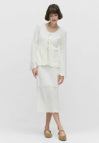 Ensemble en tricot blanc composé d'un haut à lacets à manches évasées et d'une jupe midi avec des motifs zigzag et ajourés. Chaussures plates.