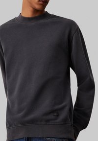 Sudadera gris oscuro hecha de un tejido suave, con cuello redondo, puños acanalados y una pequeña etiqueta con el logo cerca del dobladillo.