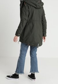 Zelená parka s kapucí, stažená v pase a se šňůrkami. Doplněná o modré džíny s roztřepeným lemem a černé tenisky.