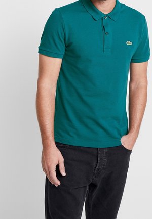 Mand iført en turkis kortærmet polo med krave og logo på venstre bryst, kombineret med sorte bukser, hånd i lommen.