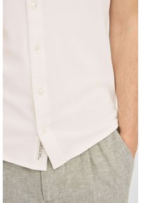 Hvit button-up skjorte med en jevn tekstur, med en klassisk krage, en knappestolpe og et merke på siden; sammen med lysegrå bukser.
