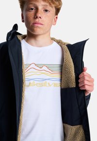Marineblaues Jackett mit einem beigen Kunstfellfutter, geöffnet, um ein weißes Grafik-T-Shirt mit bunten Bergdesigns und Text darunter zu zeigen.