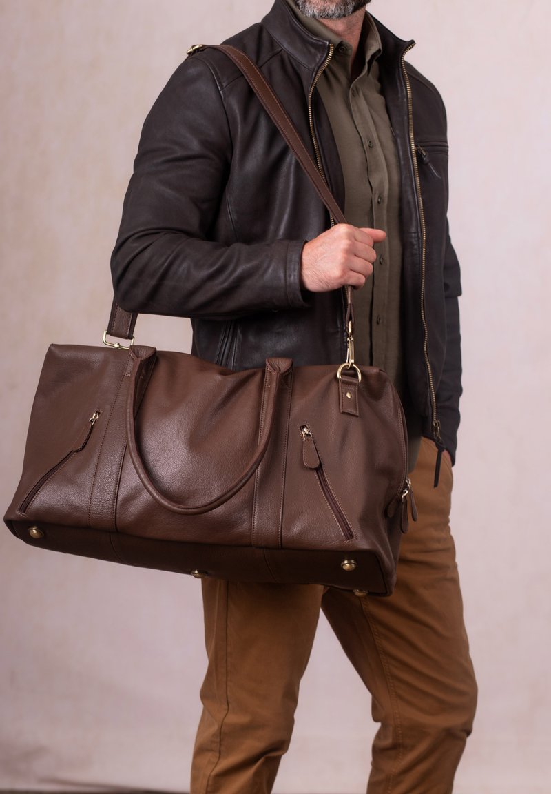 Brown leather duffel bag with a smooth texture, dual handles, and gold hardware accents. Features side pockets and a spacious design.