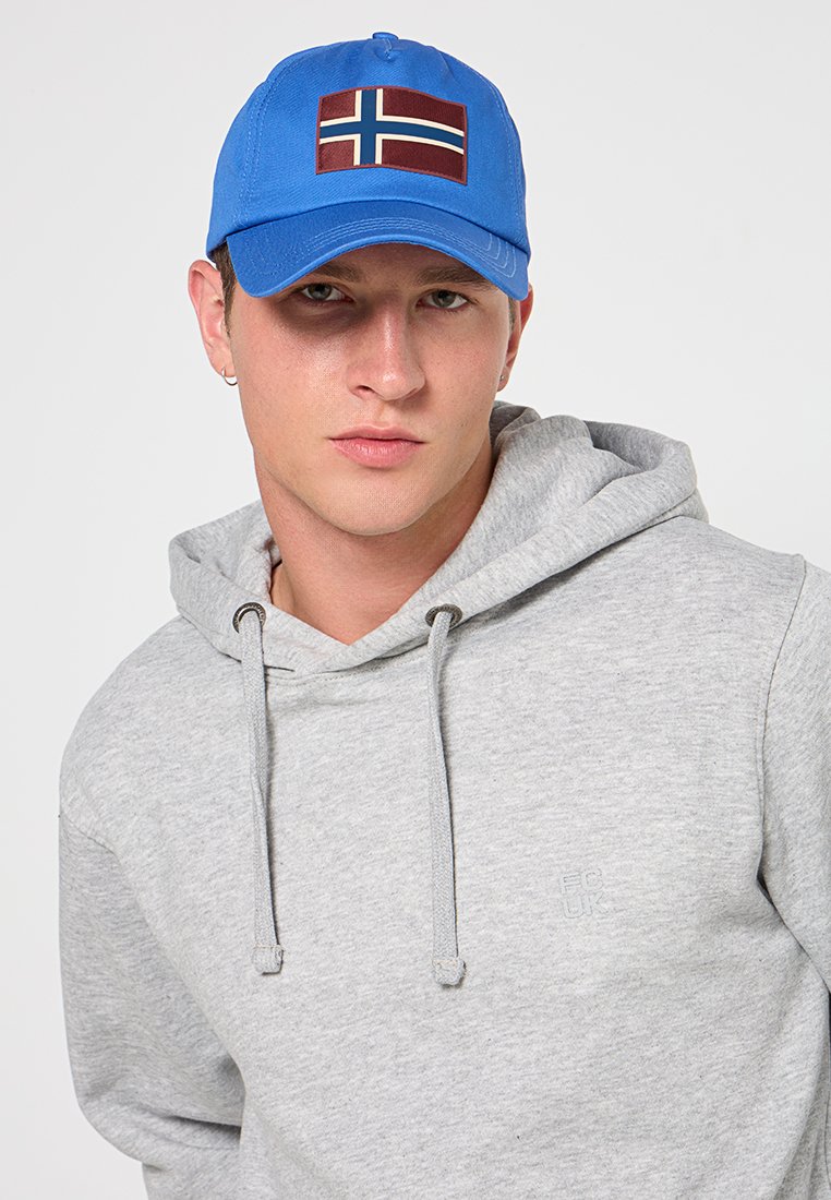 Młody mężczyzna noszący niebieską czapkę z flagą Norwegii i jasnoszary hoodies z troczkami, na jednolitym białym tle.