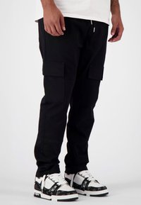 Zwarte cargobroek met twee zijzakken en elastische boorden, gemaakt van stevig stof. Gekleed met zwart-witte sneakers met een patroon.