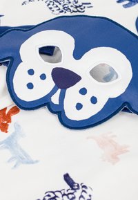 Masque pour les yeux en tissu bleu en forme de visage de chien, avec des accents blancs, de grands yeux et des motifs à pois, posé sur un fond à motifs.