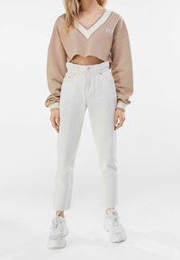 Sudadera marrón recortada con escote en V y acentos blancos, combinada con jeans de pierna recta blancos de cintura alta y zapatillas blancas.