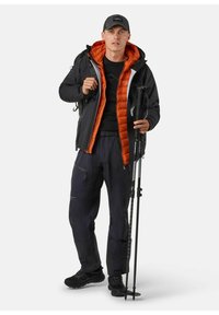 Zwarte waterdichte jas met oranje isolatie, gecombineerd met donkere broek en wandelschoenen. Houdt trekkingstokken vast, draagt een zwarte pet.