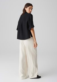 Maglietta nera a maniche corte con texture e risvolti, abbinata a pantaloni bianchi larghi e comodi e scarpe nere, vista da dietro.
