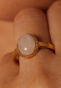 Goldring mit einem großen, ovalen, hellrosafarbenen Stein, umgeben von einem perlenbesetzten Fassungsring, der auf einem Finger mit strukturiertem Hauthintergrund liegt.