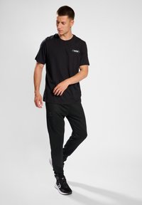 Schwarzes T-Shirt mit kurzen Ärmeln und einem Logo auf der Brust, kombiniert mit schwarzen schmal geschnittenen Sweatpants und schwarzen Sportschuhen. Einfaches Design, glatte Textur.