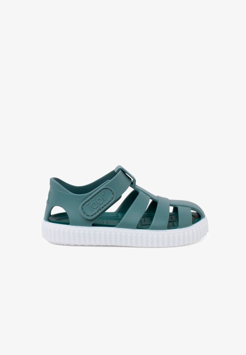 Teal rubber sandal dengan desain terbuka, tali yang dapat disesuaikan, dan sol tekstur putih. Memiliki aksen yang ditinggikan dan detail potongan di sisi-sisi.