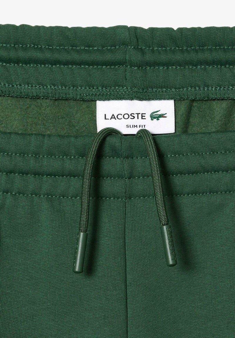 Zielone dresowe spodnie slim fit z elastycznym paskiem i teksturowanym materiałem. Posiadają metkę z logo Lacoste na pasku.