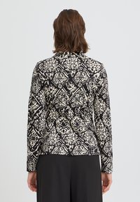 Haut à manches longues avec un motif floral noir et crème. Présente une coupe ajustée, une texture lisse et un col à revers.