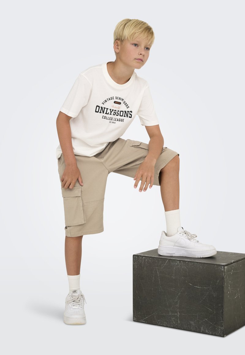 Jaunas žmogus, apsirengęs baltu medvilniniu marškinėliu su tamsiu spaudiniu, rudomis cargo šortais ir baltais sportbačiais, stovi ant juodo kvadratinio pjedestalo.