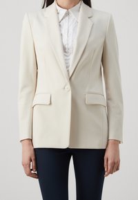 Blazer crema con solapas de pico, dos bolsillos frontales, cierre de un solo botón y una silueta entallada. Combinado con una blusa blanca.