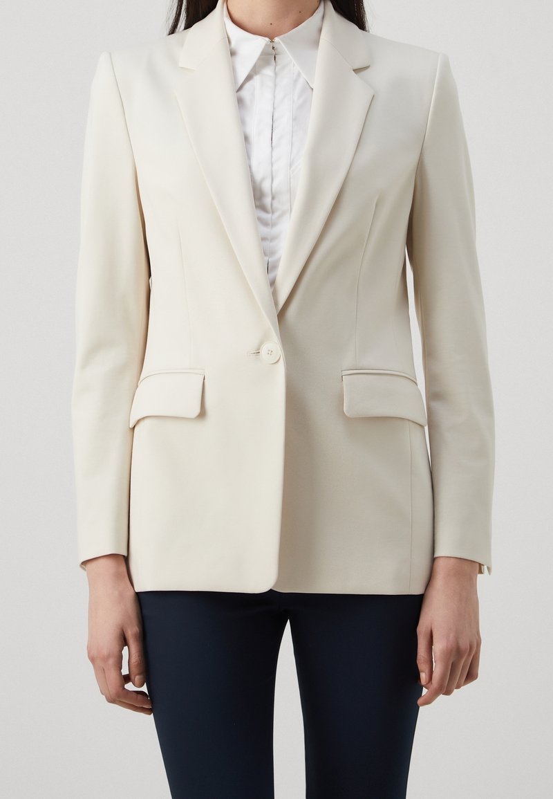 Blazer crema con solapas de pico, dos bolsillos frontales, cierre de un solo botón y una silueta entallada. Combinado con una blusa blanca.