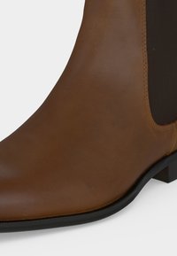 Bottine en cuir marron avec panneau latéral élastique noir et semelle noire, vue de côté en gros plan.