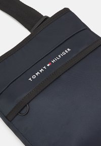 Marineblaue Nylon-Tasche mit strukturiertem Oberflächenfinish, ausgestattet mit einem Tragegurt und dem weißen "TOMMY HILFIGER"-Logo, ergänzt durch ein rotes und blaues Emblem.