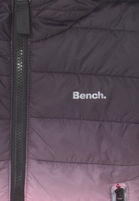 Wypełniona kurtka w odcieniach fioletu z poziomymi pikowaniami. Posiada zamek błyskawiczny i białe logo "Bench.". Gładka tekstura.