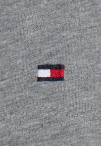 Lille rektangulær broderet logo med hvide og røde sektioner kantet i sort på gråstrikket stof.