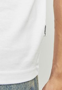 Camiseta de algodón blanca con cuello redondo y mangas cortas, con una etiqueta lateral que dice "ORIGINALS™" cerca del dobladillo. Llevada con jeans de mezclilla.