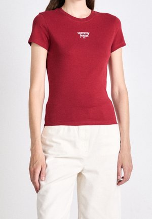Vrouw draagt een nauwsluitend rood Tommy Jeans T-shirt met korte mouwen en witte broek met hoge taille, staand tegen een effen achtergrond.