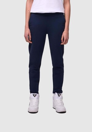 Persona che indossa pantaloni slim fit blu navy e sneakers alte bianche, in piedi con le mani rilassate ai lati, su sfondo grigio chiaro.